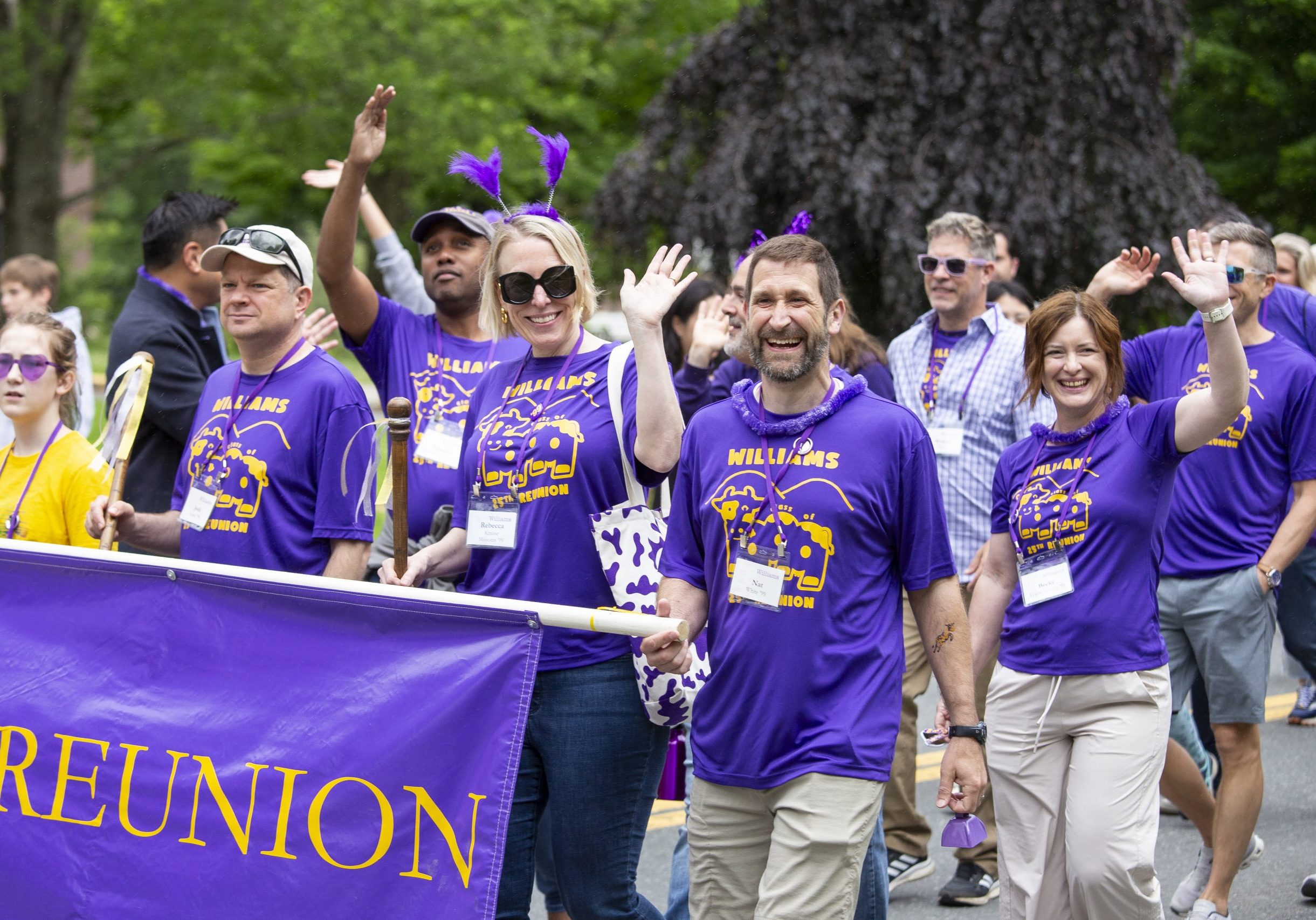 A group of people in purple shirts smile as they hold and Reunion banner and walk in a parade during their college reunion weekend.